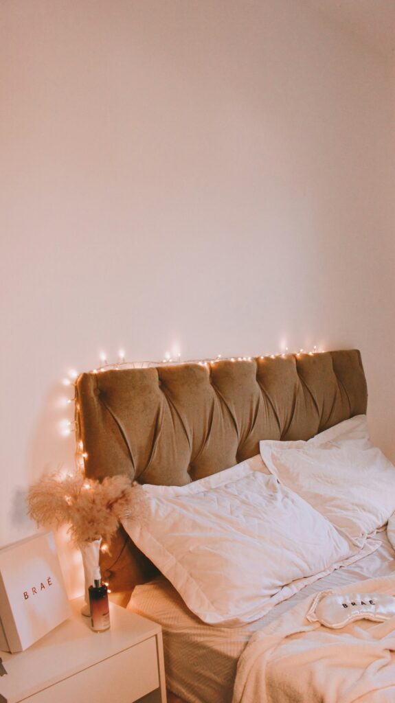 Cama con sábanas blancas, cabecero tapizado marrón y guirnalda de luces